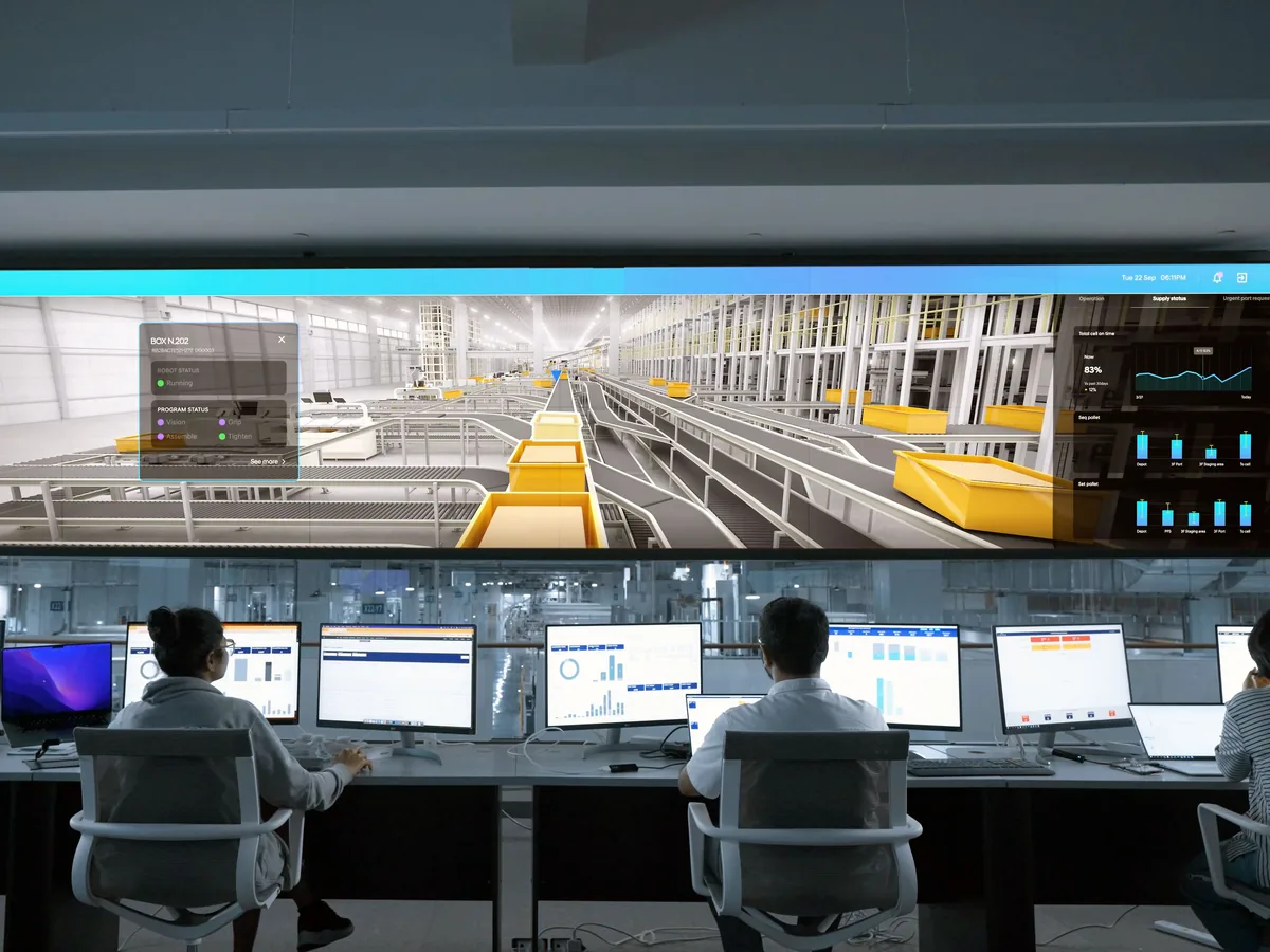Operators seen from behind in a control room, a wall screen showing a 3D view of a conveyor line