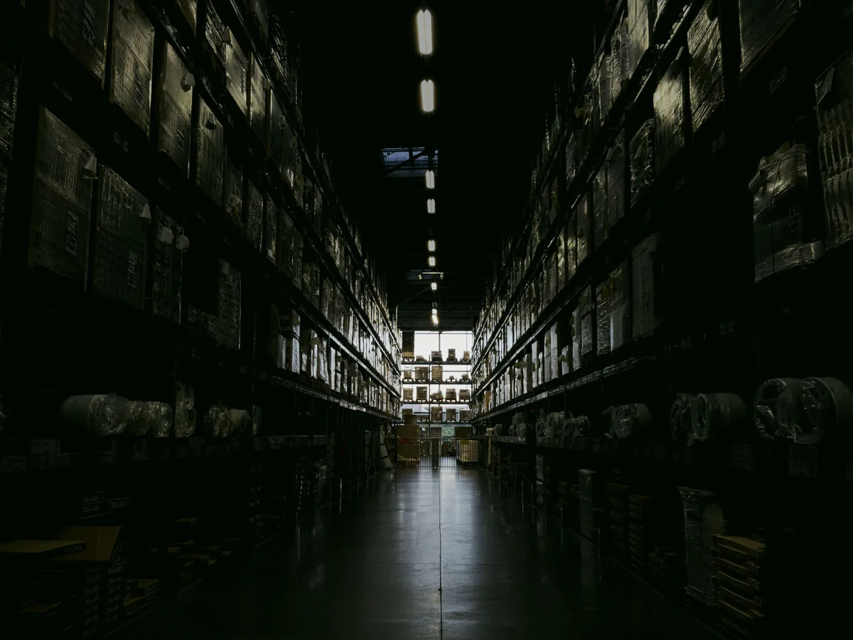A long warehouse aisle in near darkness, racks of stock stacked to the ceiling on both sides